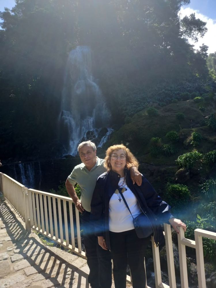 A couple standing on a bridge in front of a waterfall, small in the frame and lit from behind